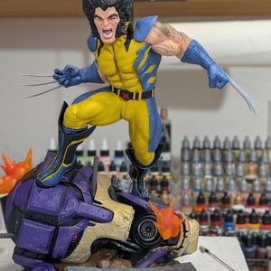 May include: A Wolverine action figure in a yellow and blue suit with extended metal claws, standing on a defeated purple and gold robot head. The figure has a snarling expression and black hair. A logo for Paynestream Studios is visible.