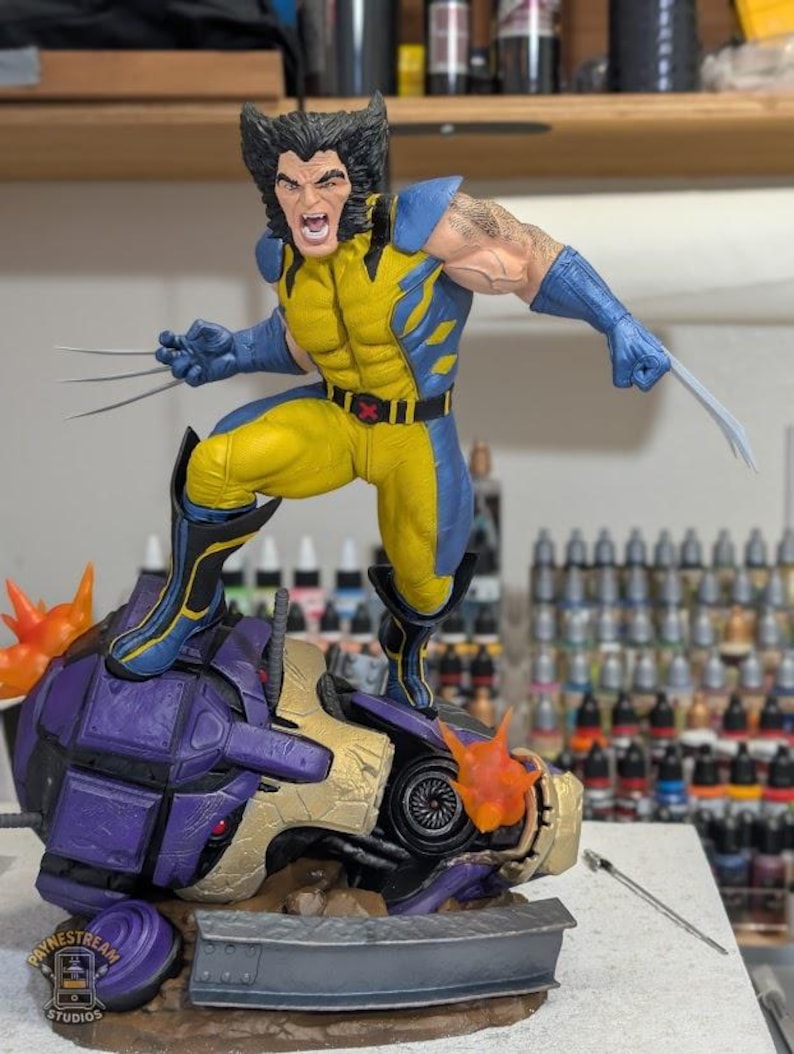 May include: A Wolverine action figure in a yellow and blue suit with extended metal claws, standing on a defeated purple and gold robot head. The figure has a snarling expression and black hair. A logo for Paynestream Studios is visible.