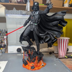 May include: A Darth Vader figurine standing on a rocky base with orange and red lava details. The figure wears a black suit and flowing cape, holding a red lightsaber in one hand with the other hand outstretched. The base features painted lava.
