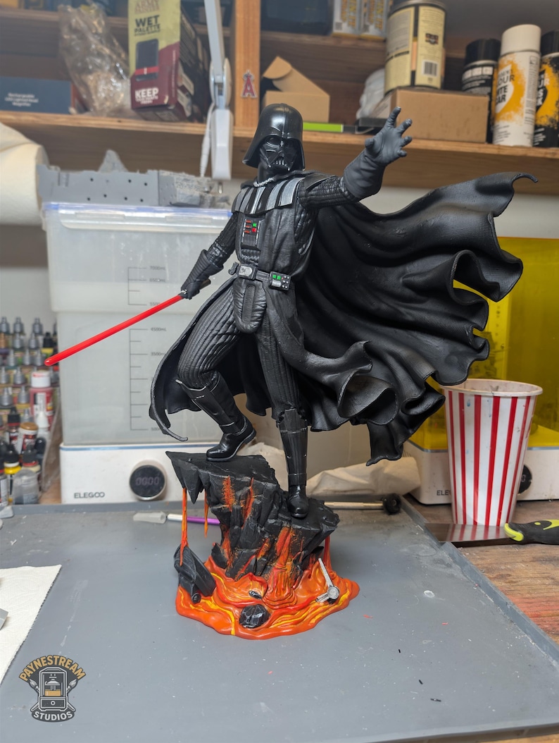 May include: A Darth Vader figurine standing on a rocky base with orange and red lava details. The figure wears a black suit and flowing cape, holding a red lightsaber in one hand with the other hand outstretched. The base features painted lava.