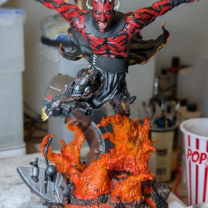 May include: A detailed Darth Maul action figure featuring red and black skin markings, a horned head, and a black outfit. The figure holds a double-bladed red lightsaber above its head, leaping from a base of sculpted orange and black lava flames.
