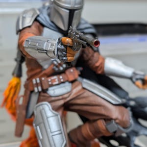 K&ouml;nnte beinhalten: Detaillierte Figur von The Mandalorian, in silberner R&uuml;stung und Helm, mit einer Blasterpistole. Die Figur ist dynamisch posiert, mit braunen und orangefarbenen Akzenten auf der Kleidung. Der Sockel hat ein feuriges orange-gelbes Design.