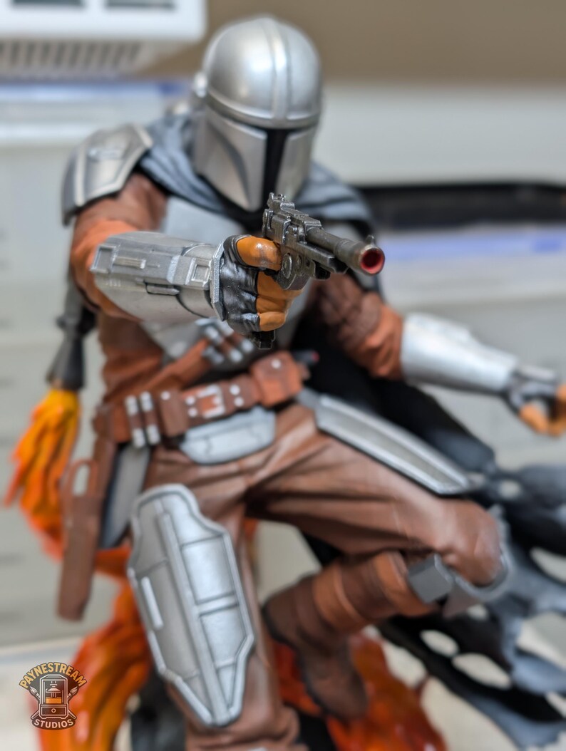 K&ouml;nnte beinhalten: Detaillierte Figur von The Mandalorian, in silberner R&uuml;stung und Helm, mit einer Blasterpistole. Die Figur ist dynamisch posiert, mit braunen und orangefarbenen Akzenten auf der Kleidung. Der Sockel hat ein feuriges orange-gelbes Design.