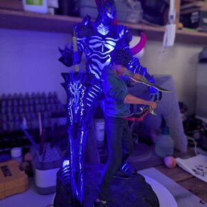 May include: A figurine featuring a young person in a blue hoodie and dark pants holding two daggers, standing in front of a large, glowing blue and black armored entity with a red horn and a massive, illuminated sword on a dark, rocky base.