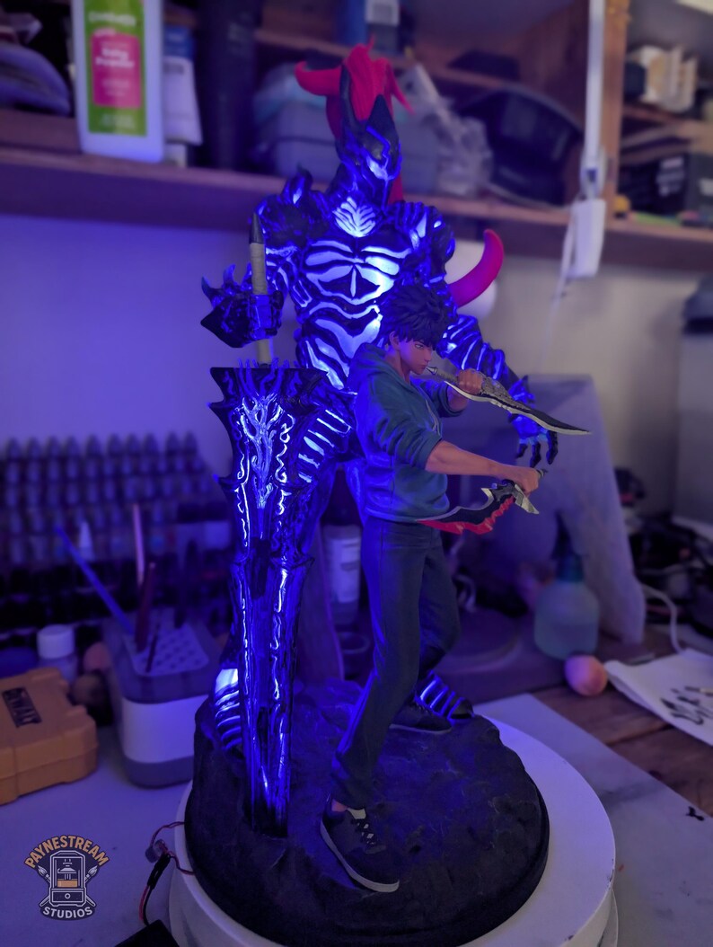 May include: A figurine featuring a young person in a blue hoodie and dark pants holding two daggers, standing in front of a large, glowing blue and black armored entity with a red horn and a massive, illuminated sword on a dark, rocky base.