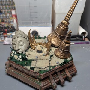 May include: A diorama featuring a grey stone Buddha head sculpture, two gold-colored temple bells, and a tall gold spire on a cracked stone base with green moss and small white flowers. The base is constructed from brown wooden beams.