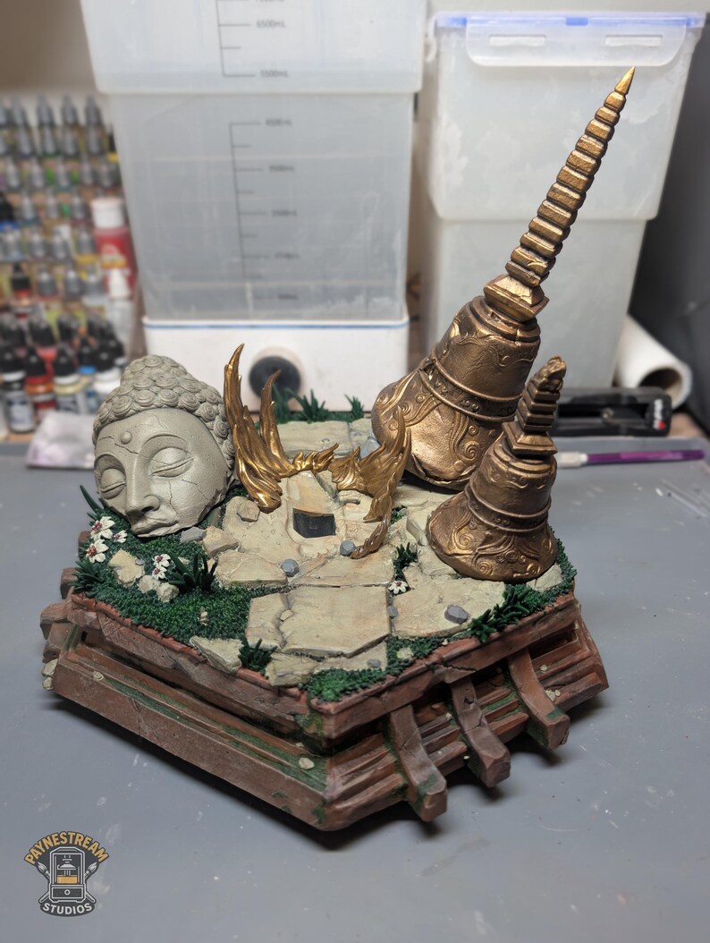 May include: A diorama featuring a grey stone Buddha head sculpture, two gold-colored temple bells, and a tall gold spire on a cracked stone base with green moss and small white flowers. The base is constructed from brown wooden beams.