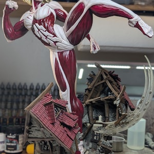 May include: A detailed figurine of a blonde humanoid giant with exposed red muscle tissue and white skin patches, captured in a dynamic kicking pose. The figure stands atop a miniature, ruined timber-framed house with a red tiled roof and debris.