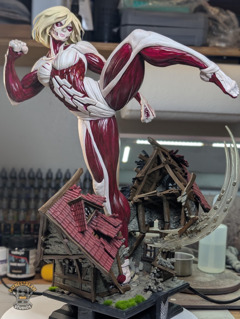 May include: A detailed figurine of a blonde humanoid giant with exposed red muscle tissue and white skin patches, captured in a dynamic kicking pose. The figure stands atop a miniature, ruined timber-framed house with a red tiled roof and debris.
