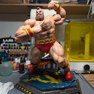 May include: A muscular, bearded figurine with a mohawk, wearing red briefs, red wristbands with yellow stripes, and red boots. The figure stands on a hexagonal base featuring a yellow hammer and sickle emblem and a metal chain.