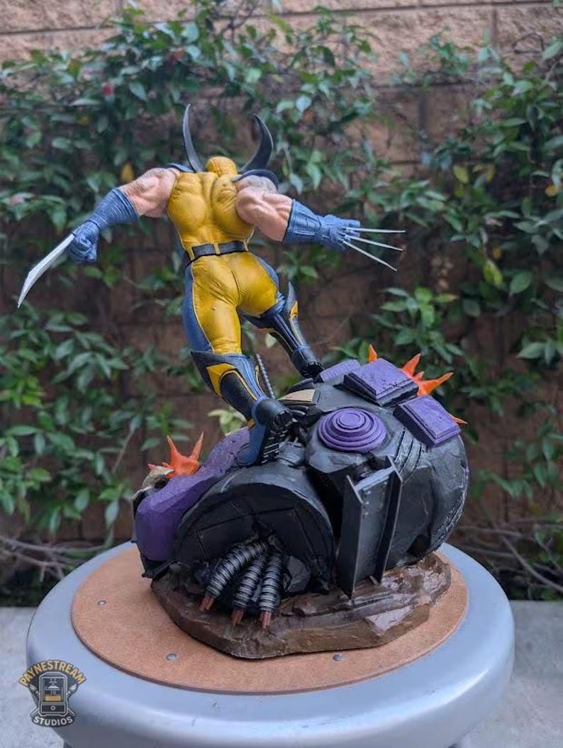 May include: A Wolverine action figure in a dynamic pose, clad in a yellow and blue costume, with claws extended. The figure stands atop a detailed, dark-colored base with purple and orange accents. The base has the text "PAYNESTREAM STUDIOS".