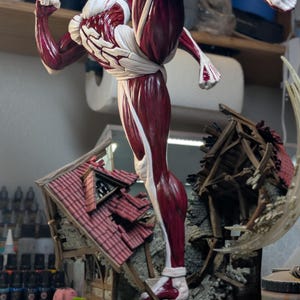 May include: A detailed figurine of a blonde character with exposed red muscle tissue and white skin, captured in a dynamic high-kick pose. The figure stands atop a base featuring a miniature, partially destroyed house with a red tiled roof.