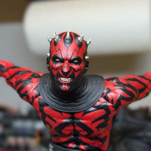 K&ouml;nnte beinhalten: Eine detaillierte Figur mit rotem und schwarzem Hautmuster, gelben Augen und kleinen H&ouml;rnern auf dem Kopf. Die Figur hat einen grimmigen Gesichtsausdruck mit sichtbaren Z&auml;hnen und tr&auml;gt einen schwarzen Kragen sowie einen schwarzen G&uuml;rtel.
