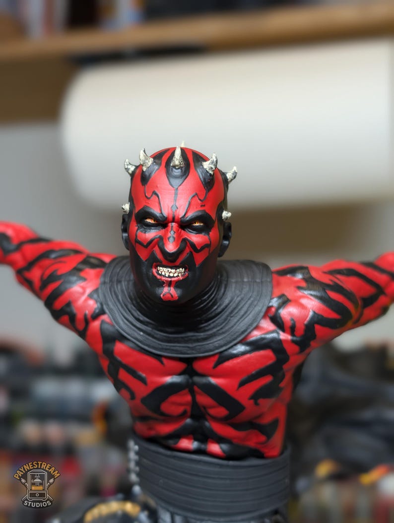 K&ouml;nnte beinhalten: Eine detaillierte Figur mit rotem und schwarzem Hautmuster, gelben Augen und kleinen H&ouml;rnern auf dem Kopf. Die Figur hat einen grimmigen Gesichtsausdruck mit sichtbaren Z&auml;hnen und tr&auml;gt einen schwarzen Kragen sowie einen schwarzen G&uuml;rtel.