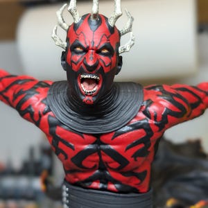 K&ouml;nnte beinhalten: Eine detaillierte Figur mit rot-schwarzem Hautmuster, gelben Augen und spitzen Z&auml;hnen, die eine Krone aus spitzen H&ouml;rnern auf dem Kopf tr&auml;gt. Die Figur tr&auml;gt einen dunklen Kragen und einen schwarzen G&uuml;rtel und zeigt eine dynamische Pose.