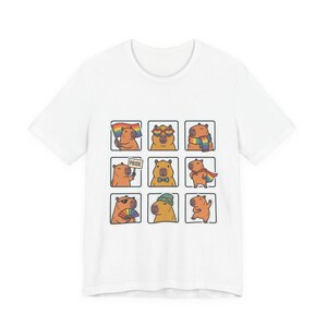 May include: White t-shirt featuring a grid of nine cartoon capybaras, each with a unique accessory or prop. Some capybaras hold a rainbow flag, wear sunglasses, or sport a bow tie. The word "PRIDE" is on a sign.