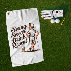 May include: White golf towel with the text "Swing, Swear, Drink, Repeat" and a cartoon golfer. A golf glove, two golf balls, and a tee are on the green grass.