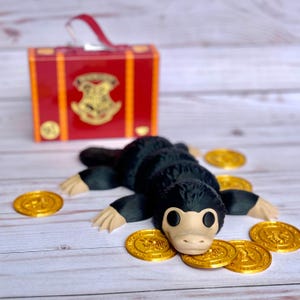May include: A black and brown Niffler figurine with a long snout and beady eyes, surrounded by gold coins. The figurine is sitting on a white wooden surface. A red suitcase with gold accents and a Hogwarts crest is in the background.
