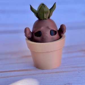 May include: A small brown and green ceramic figurine of a plant with a face, in a beige pot. The figurine has a sad expression.