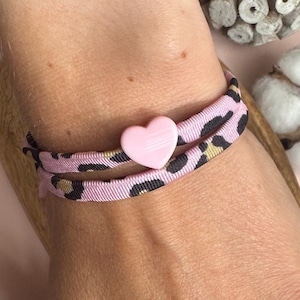 Könnte beinhalten: Ein doppelt gewickeltes elastisches Haarband mit hellrosa Hintergrund und einem schwarzen sowie goldenen Leopardenmuster. Das Haaraccessoire ist mit einem kleinen, glatten, einfarbigen hellrosa herzförmigen Anhänger in der Mitte verziert.
