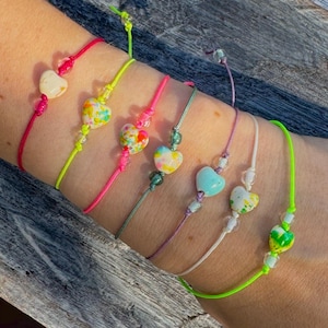 Könnte beinhalten: Eine Auswahl an Armbändern mit herzförmigen, farbigen Perlen. Jedes Armband hat eine Schnur in einer anderen Farbe, darunter Rot, Grün, Pink und Lila. Die Herzperlen sind in verschiedenen Farben und Mustern gehalten.