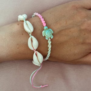 Könnte beinhalten: Ein handgefertigtes Armband mit weißen Muscheln und einem türkisfarbenen Schildkrötenanhänger. Das Armband ist mit rosa, blauen und weißen Kordeln versehen. Das Armband ist für das Handgelenk konzipiert.