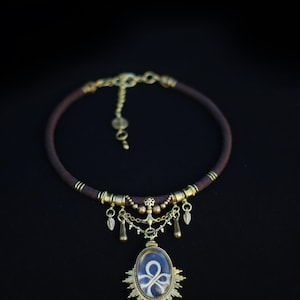 Peut inclure: Un collier ras-de-cou en cordon marron avec des ornements en métal couleur laiton antique, comprenant des perles cylindriques, de petites breloques en forme de feuille et un pendentif ovale central avec un cadre rayonnant entourant un nœud blanc.