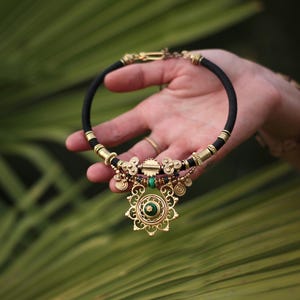 Peut inclure: Un collier ras-du-cou en cordon noir avec des accents en métal doré et un pendentif circulaire central incrusté d'une pierre de malachite verte. Le design comprend des détails en filigrane, des breloques pendantes et des perles cylindriques dorées.