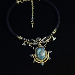Peut inclure: Un collier en cordon noir avec une grande pierre labradorite rectangulaire aux reflets bleu-gris, montée sur une structure en laiton. Le pendentif est orné de petites perles rondes, de fil de laiton et de deux breloques en forme de hache.