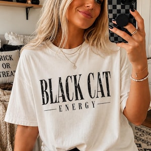 May include: An off-white short-sleeve t-shirt featuring the black text "BLACK CAT ENERGY" printed across the chest in a distressed serif font. The shirt has a crew neckline and a relaxed fit, worn with a layered gold necklace and a beaded bracelet.