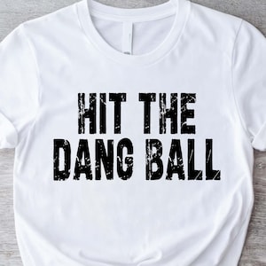 May include: White t-shirt with the phrase "HIT THE DANG BALL" printed in distressed black letters. The shirt is made of a soft material and has short sleeves. The shirt is knotted at the waist.
