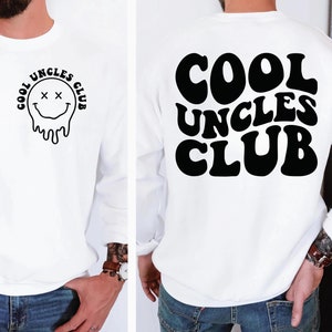 Puede incluir: Sudadera blanca con una cara sonriente negra que gotea y el texto "Cool Uncles Club" en la parte delantera. La parte posterior de la sudadera tiene el texto "Cool Uncles Club" en letras negras grandes.