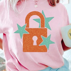 May include: Pale pink t-shirt featuring an orange glitter padlock design and three teal stars. A white mug with a yellow smiley face is held. The shirt is a casual style.