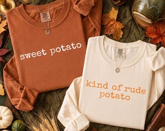 Puede incluir: Una camiseta de manga larga color óxido con el texto blanco sweet potato y una camiseta de manga larga color crema con el texto naranja kind of rude potato. Ambas prendas tienen cuello redondo y están sobre una tela verde oscuro.