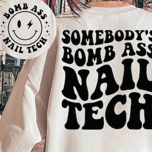 May include: White sweatshirt with black text that reads "Somebody's Bomb Ass Nail Tech". A smaller black and white sticker with a smiley face and the text "Bomb Ass Nail Tech" is on the left side of the sweatshirt.