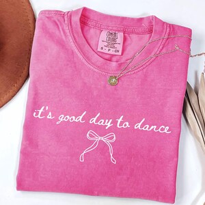 May include: A bright pink short-sleeved t-shirt with a crew neckline. The shirt features the white text "it's good day to dance" printed above a simple white line drawing of a bow. A gold-toned necklace with a small circular pendant rests on the shirt.