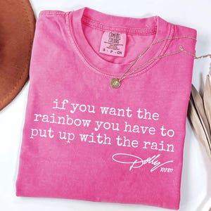 Pode incluir: Uma t-shirt de manga curta cor-de-rosa brilhante com texto branco que diz: if you want the rainbow you have to put up with the rain, seguido de uma assinatura Dolly XOXO. A t-shirt tem uma etiqueta da marca Comfort Colors no pescoço.