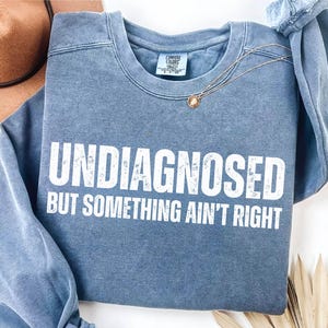 Może przedstawiać: Niebieska bluza z okrągłym dekoltem i postarzanym białym nadrukiem graficznym o treści UNDIAGNOSED BUT SOMETHING AIN'T RIGHT. Odzież posiada prążkowany kołnierzyk i długie rękawy oraz metkę marki Comfort Colors widoczną przy wewnętrznym dekolcie.