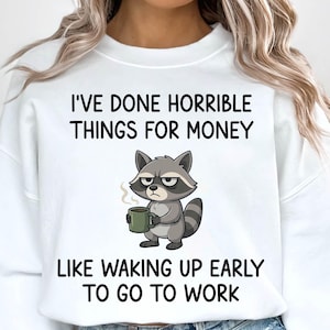 May include: A white crewneck sweatshirt featuring a cartoon raccoon holding a steaming mug. The text reads: I'VE DONE HORRIBLE THINGS FOR MONEY LIKE WAKING UP EARLY TO GO TO WORK. The raccoon has grey fur, a striped tail, and a grumpy facial expression.