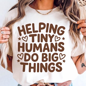 Puede incluir: Camiseta color crema con la frase marrón "HELPING TINY HUMANS DO BIG THINGS", decorada con corazones y estrellas. La camiseta está hecha de un material suave.