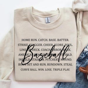 May include: A beige baseball-themed sweatshirt with black text. The word "Baseball" is written in a cursive font. The sweatshirt has a crew neck and long sleeves. The text includes baseball terms like "Home Run" and "Strike".