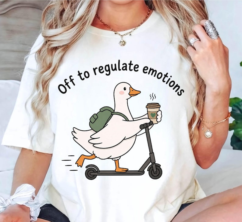 May include: A white t-shirt featuring a graphic of a white duck with an orange beak and feet, wearing a green backpack, riding a black scooter, and holding a coffee cup. The text above the duck reads, Off to regulate emotions.