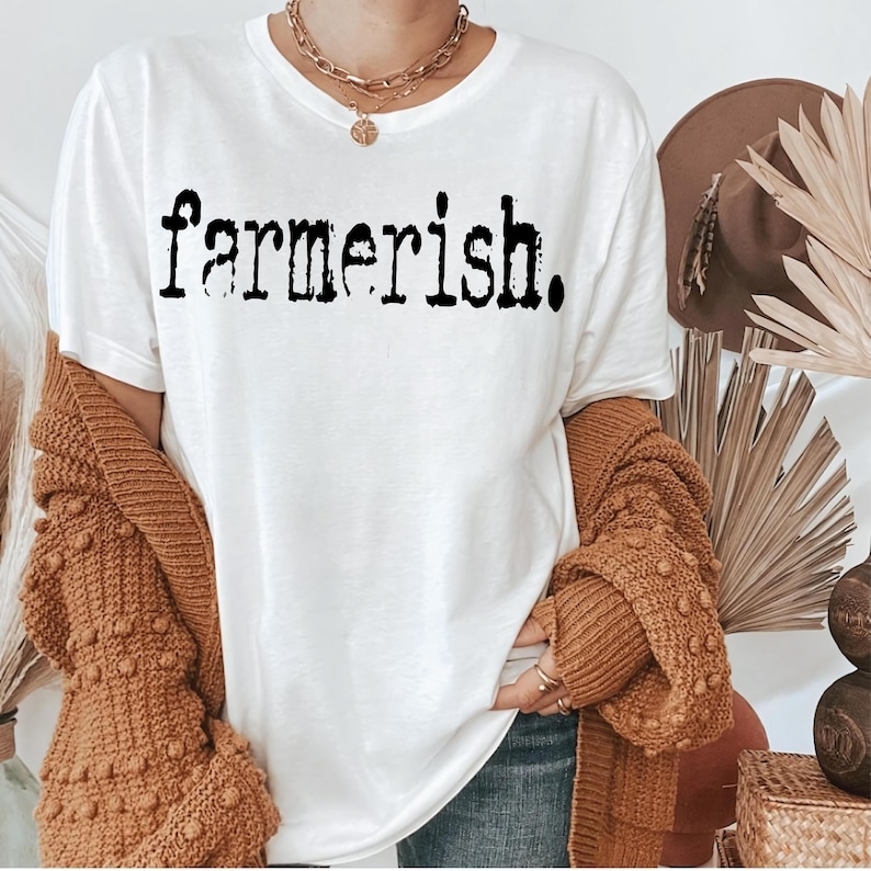 Op de afbeelding: Wit T-shirt met de tekst "farmerish." in zwart gedrukt. Het shirt wordt gedragen door een persoon met een bruine cardigan en jeans.