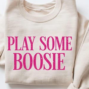May include: A beige sweatshirt with the words "PLAY SOME BOOSIE" printed in bright pink, distressed lettering. The sweatshirt has a crew neck and long sleeves.