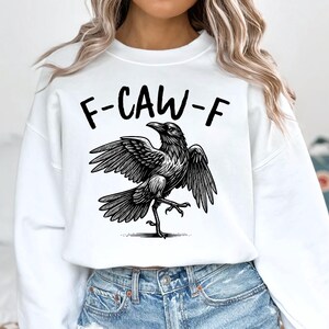 May include: White sweatshirt with a black graphic of a crow with outstretched wings and the text "F-CAW-F" above it. The crow is detailed with shading, giving it a realistic appearance. The sweatshirt is paired with blue denim jeans.