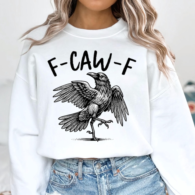 May include: White sweatshirt with a black graphic of a crow with outstretched wings and the text "F-CAW-F" above it. The crow is detailed with shading, giving it a realistic appearance. The sweatshirt is paired with blue denim jeans.