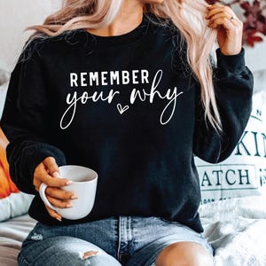 Puede incluir: Sudadera negra con el texto "Remember your why" en letras blancas con un pequeño símbolo de corazón. La sudadera la lleva puesta una persona con el pelo largo y rubio.