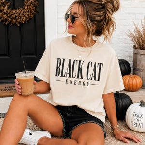 May include: A cream-colored short-sleeve t-shirt with the text BLACK CAT ENERGY printed in black on the front. The shirt features a crew neckline and a relaxed fit, paired with black denim shorts and white canvas sneakers.