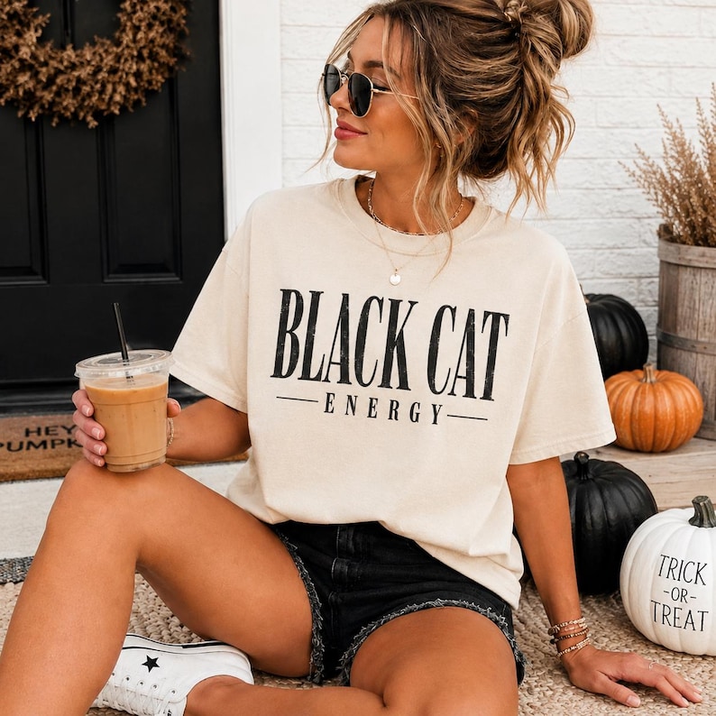 May include: A cream-colored short-sleeve t-shirt with the text BLACK CAT ENERGY printed in black on the front. The shirt features a crew neckline and a relaxed fit, paired with black denim shorts and white canvas sneakers.