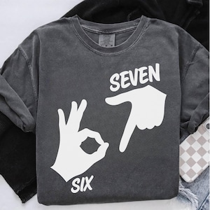 May include: Dark gray long-sleeve shirt with a white graphic design. The design features the word "SEVEN" above a hand pointing down and the word "SIX" below a hand making an "OK" gesture.
